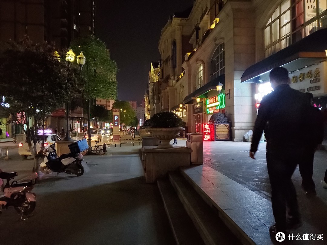 较暗的商业街道路夜景，画面亮度真实，树叶细节丢失明显